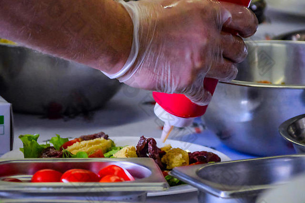 在烹饪大师课上，主厨用不锈钢碗将红色瓶子里的酱汁滴在准备好的盘子上