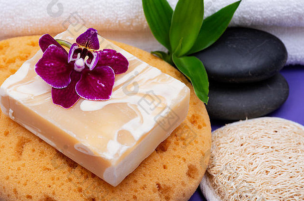 Spa健康概念。杏仁山羊奶皂、泡沫沐浴海绵、玄武岩石材、天然丝瓜海绵、紫色背景上的竹子和兰花