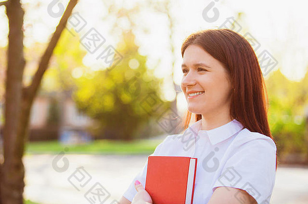 在阳光明媚的背景下，兴奋的微笑着的女学生在户外拿着一本红皮书。校园里拿着一本书微笑着的年轻女学生。大学校园