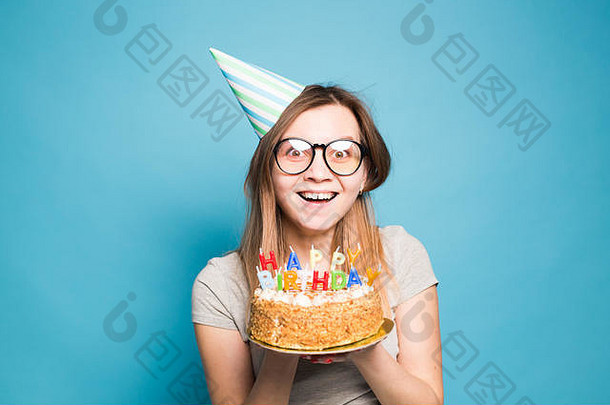 特写镜头：戴着眼镜、戴着问候纸帽子、手拿生日蛋糕、站在蓝色背景上的滑稽乐观的女孩。