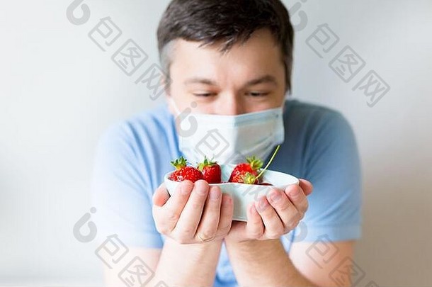 特写镜头：戴着医用口罩的年轻人拿着盛有草莓的碗。戴着防毒面具的男人。从冠状病毒中恢复。停止COVID-19大流行