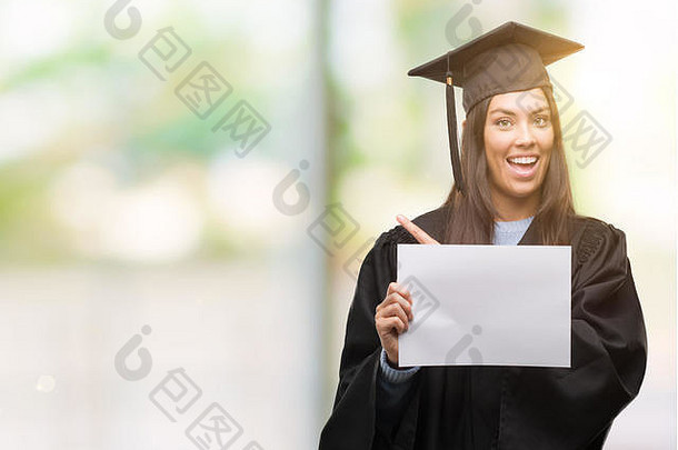 年轻的拉美裔女人穿毕业统一的持有文凭纸快乐指出手手指一边