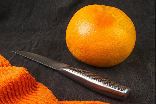 一个大葡萄柚，带一把刀和一张橙色餐巾，背景为黑色织物