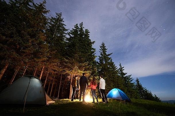 后视图一群游客站在帐篷附近的熊熊大火旁，在星空下的松林背景下。夜空下远处可见的小山。