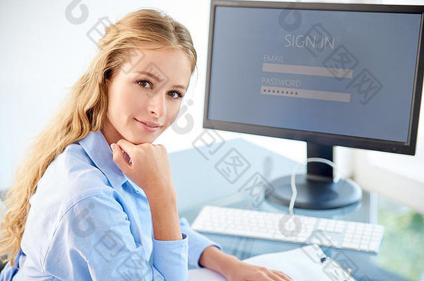 一张年轻貌美的女商人坐在电脑前看着相机微笑的照片。