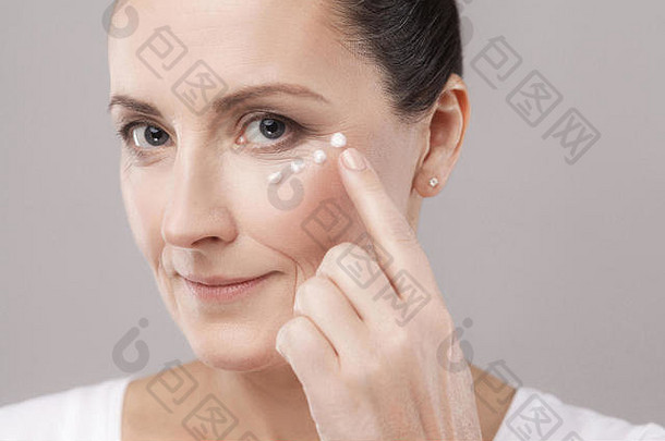 中间岁的女人完美的脸皮肤应用化妆品奶油手指皮肤眼睛相机孤立的灰色背景情郎