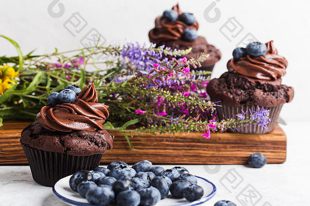 白色桌子上的巧克力纸杯蛋糕，奶油和蓝莓，野花花束，特写镜头，美味的节日甜点