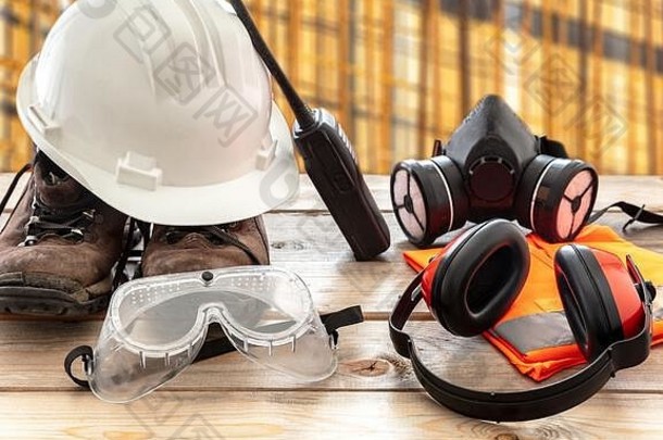 安全生产防护设备。木制工作台上的工业防护装备，模糊了施工现场背景。安全帽、靴子、耳罩、呼吸系统