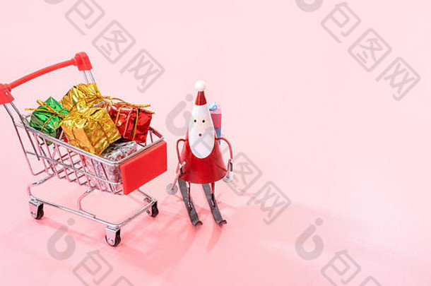 圣诞购物概念，迷你红色购物车手推车，带圣诞老人玩具和礼品盒，背景为淡粉色，空白空间，特写