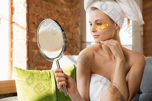 选美日。穿着毛巾在家做日常护肤的女人。戴上金色眼罩，看着镜子里的自己。美容、自我护理、化妆品、青春的概念。
