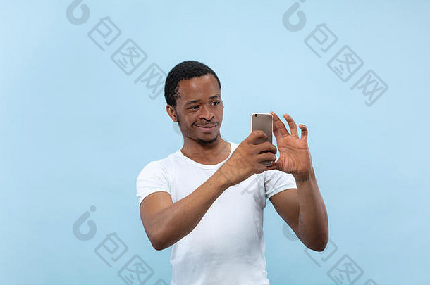 蓝色背景上穿着白衬衫的年轻非裔美国人的半身特写肖像。<strong>人类</strong>的情感，面部表情，广告概念。在他的智能<strong>手机</strong>上拍摄照片或视频<strong>日</strong>志内容。