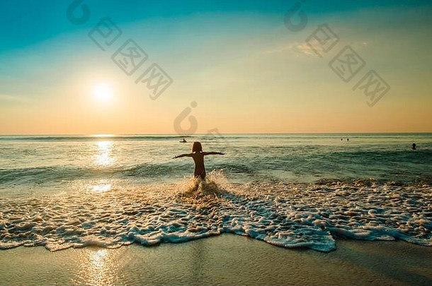 年轻女孩在清澈的海水边的热带海滩上玩耍，夕阳余晖中，蓝天在水中嬉戏，海浪<strong>来袭</strong>。