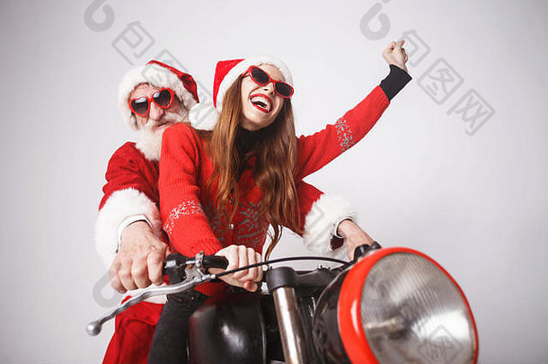快乐的年轻圣诞老人夫人戴着圣诞帽、红色毛衣和太阳镜，与圣诞老人一起骑着摩托车上高速，新年、圣诞节、节假日、纪念品、礼品、购物、折扣、商店、雪人圣诞老人、化妆、发型、狂欢节。