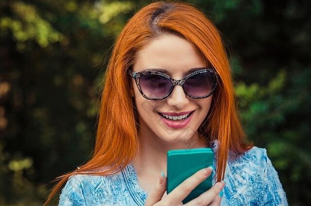 短讯服务。一位身穿蓝色牛仔裤的红头发年轻女子一边在城市公园散步一边发短信，绿色的户外背景