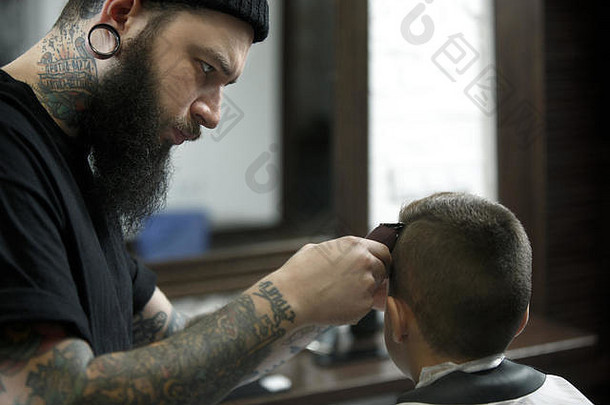 <strong>儿童</strong>理发师在黑暗的背景下给小男孩理发。心满意足的可爱学龄前男孩正在理发。大师的手上有“剃须”这个字的纹身