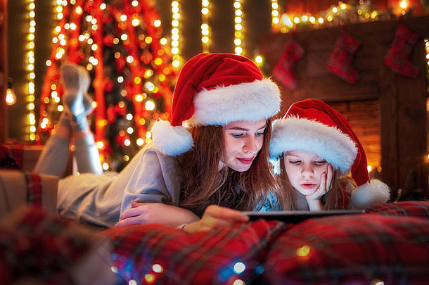 圣诞节时间。躺在床上，戴着圣诞老人帽、穿着睡衣、微笑着观看有趣视频或在数码平板电脑上挑选礼物的家庭母女