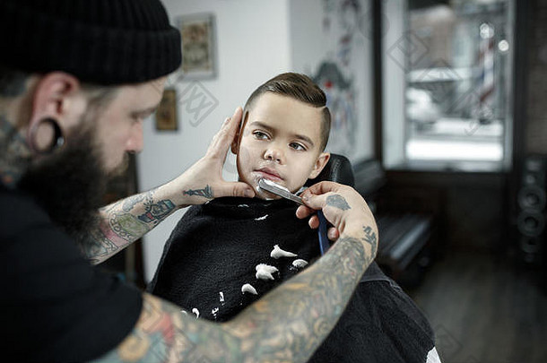 儿童理发师在黑暗的背景下给小男孩理发。心满意足的可爱学龄前男孩正在理发。大师的手上有“剃须”这个字的纹身