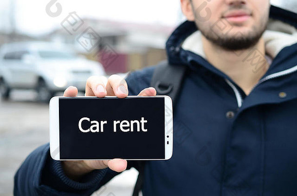 冬天，一个年轻人在一辆汽车和一幢大房子的背景上，在智能手机的显示屏上显示了一段文字。租车。