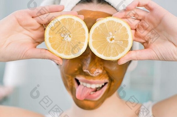 年轻漂亮的女孩，脸上戴着棕色的泥面具，手里拿着半个柠檬水果，遮住了眼睛。照顾皮肤的少女。美容治疗。