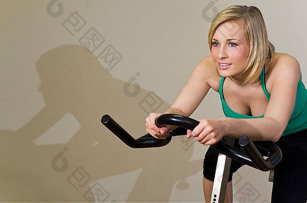 一个漂亮的年轻女子在健身房骑着健身车锻炼，她的影子就在她旁边的墙上，就像一个竞争者