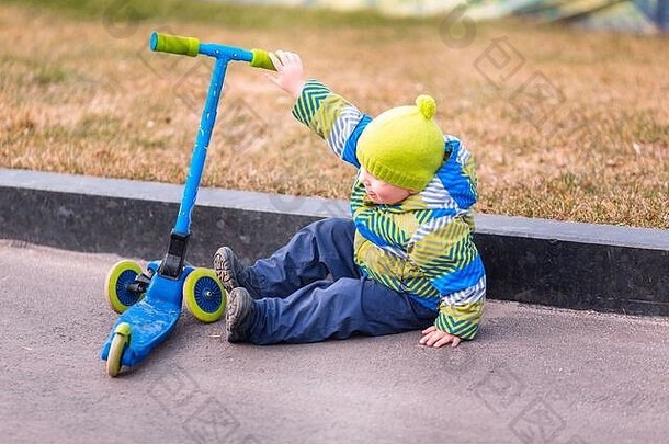 可爱的小男孩从他的滑板车上摔下来。骑脚踏车时受伤的孩子。滑板车坠落事故。男孩试图站起来