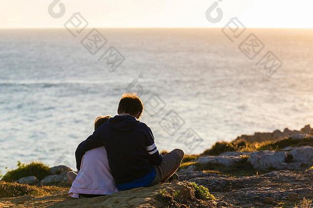 小女孩和少年男孩拥抱在一起，看着日落的大海