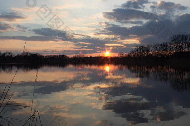 美丽的田园风光——湖面上的夕阳。夕阳照亮的云彩反射在水面上。湖边树木丛生。向左