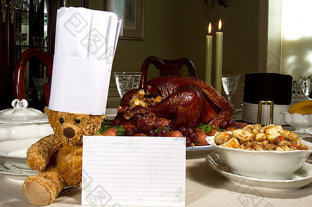 餐桌上摆满了感恩节大餐，一只泰迪熊戴着厨师帽，手里拿着一张食谱卡