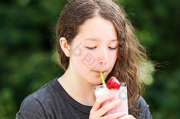 照片年轻的女孩喝着草莓奶昔夏天一天模糊明亮的绿色树背景