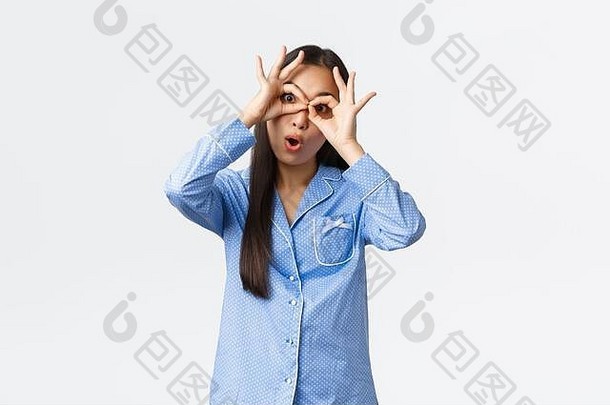 一个穿着蓝色睡衣、风趣而乐观的美丽亚洲女孩在做鬼脸，用手展示假眼镜，开玩笑地盯着摄像机，玩着嘲弄