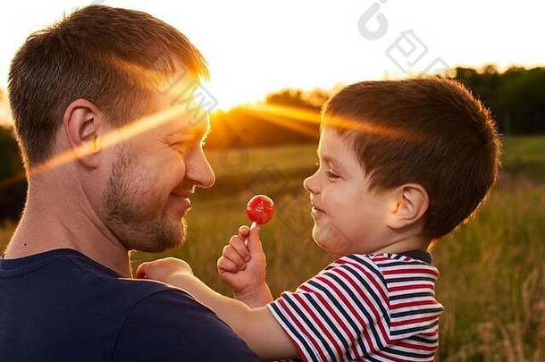 傍晚日落时分，爸爸和儿子在一片黄色的夏日田野里散步，吃着棍子上的糖果。父亲节，爱在家庭中，父亲的角色