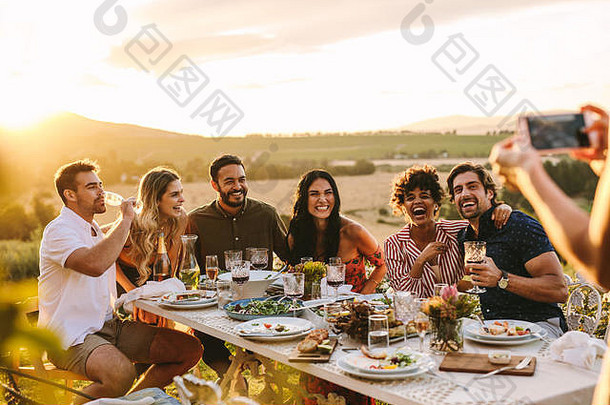 一群男女在户外晚宴上合影留念。在晚宴上，一位女士亲手为她的朋友拍照。