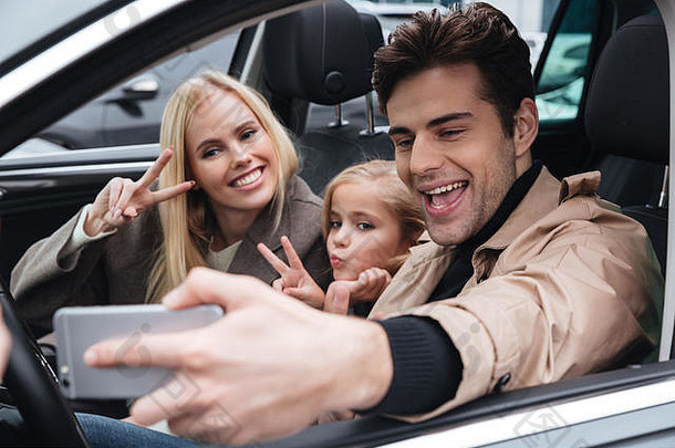 一个快乐的年轻人和他的妻子和女儿坐<strong>在</strong>车里。一边看一边用手机自拍。