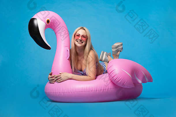 身着泳衣、戴着粉红色眼镜的金发女郎躺在蓝色背景下的粉色充气火烈鸟上