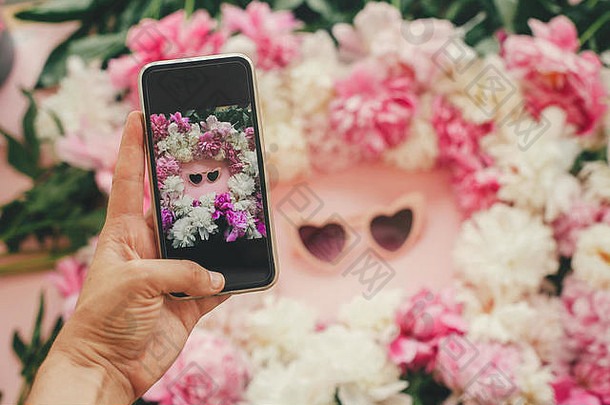 手持手机，在粉色平铺纸上，用时尚的粉色和白色牡丹花架拍摄粉色心形太阳镜。Instagram博客