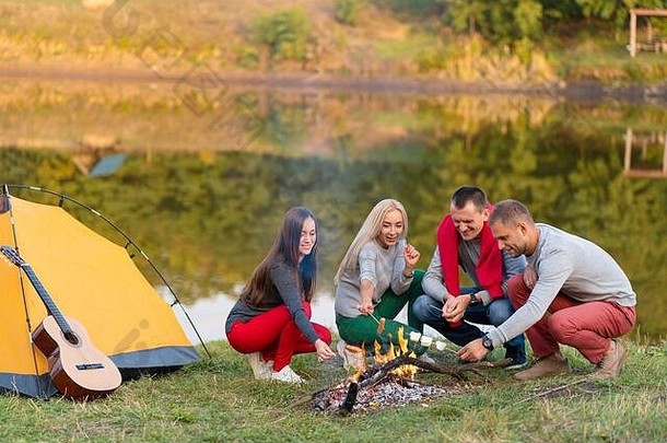 旅行，旅游，远足，野餐和人的概念-一群快乐的朋友在湖附近的篝火上炸香肠