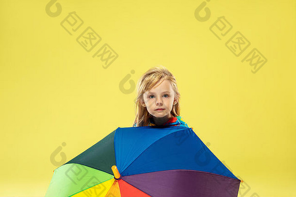 黄色摄影棚背景上，一位穿着雨衣、带彩虹色雨伞的时尚女孩的全长肖像。看起来很高兴。儿童<strong>秋</strong>季和春季时装。可爱时尚的金发女孩。