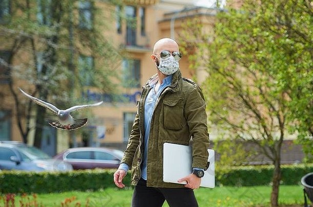 一位秃头男子戴着医用口罩，留着胡子，以避<strong>免冠</strong>状病毒的传播。他在公园里带着笔记本电脑散步。一个戴着n95面罩和飞行员的家伙