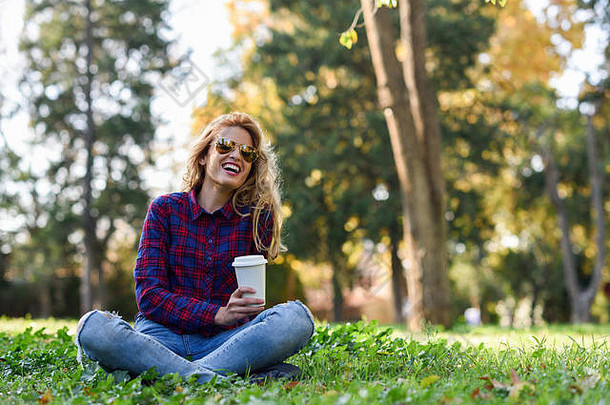 年轻快乐的女人，穿着格子衬衫和蓝色牛仔裤，带着牙齿般的微笑和太阳镜。在公园里喝咖啡的金发女孩穿着cas坐在草地上