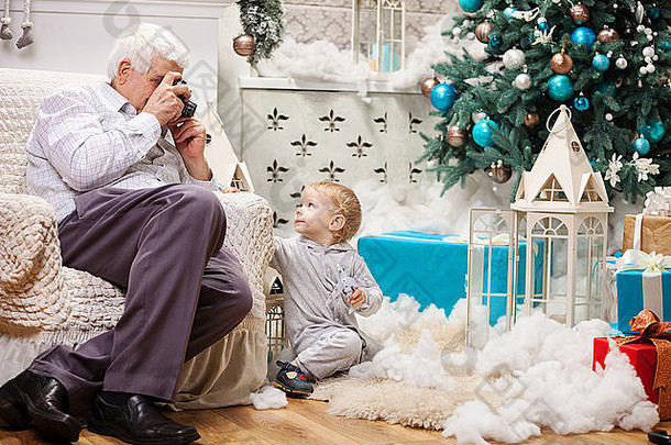 老人坐在家里的圣诞树旁给蹒跚学步的孙子拍照