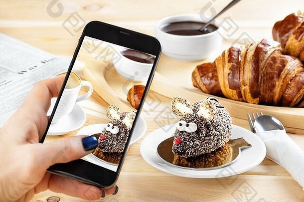 女人用手机拍下食物的照片。欧式早餐，羊角面包，鼠标形状的蛋糕和木制背景上的咖啡。Smartph
