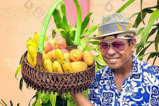 肖像微笑菲律宾男人。穿fedora他太阳镜花衬衫持有篮子新鲜的水果
