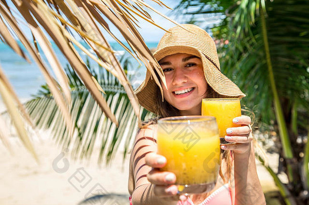 戴着夏日帽子的美丽女孩，在海滩上棕榈叶的背景下喝着新鲜的饮料，女孩提供了一种饮料、特写、度假的概念