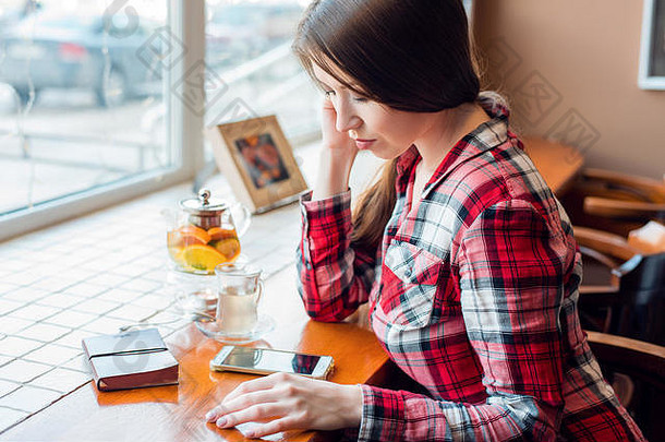 下午，在一家靠窗的咖啡馆里，一个穿着衬衫的女生看着电话，读着一条短信，茶壶里放着一杯茶，一本笔记本放在一张木桌上。