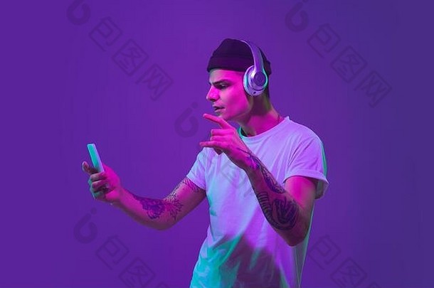 用智能手机听音乐，跳舞。白人男子的肖像画被隔离在紫色工作室背景上，粉红色霓虹灯下。漂亮的男模特。人类情感的概念，面部表情，销售，广告。