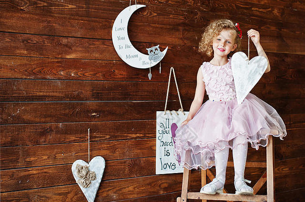 一个穿着衣服的美丽女孩的肖像，手里拿着一个玩具，坐在靠着木墙的木梯子上。