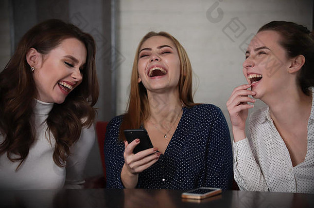三个年轻漂亮的女人坐在咖啡馆里看着电话，笑着