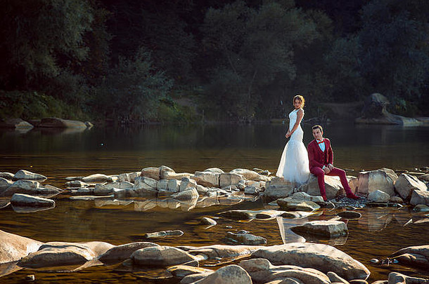 日落时分，美丽的新婚夫妇在镜头前摆姿势。迷人的红头发新娘站在河边坐着的新郎旁边。