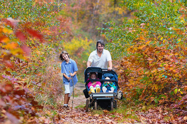 一家人在秋天公园里用婴儿车徒步旅行。活跃的父亲、婴儿和蹒跚学步的孩子坐在双人推椅上。健康的爸爸带着慢跑婴儿车和小孩走路