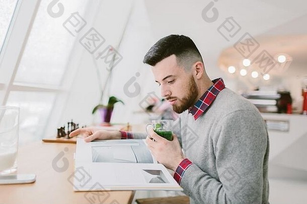 年轻的有胡子的男人。咖啡馆坐在长表格窗口的家伙持有玻璃绿色叶子手<strong>杂志</strong>男人。